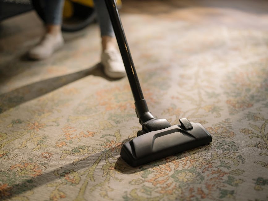 A close-up view of a person using a vacuum cleaner on a patterned carpet in a well-lit room. The vacuum cleaner has a black head with a control switch, and the person’s white shoes and part of their legs are visible in the background. The carpet features a floral design with muted colors, and the surface appears clean and well-maintained, showcasing effective surface cleaning suitable for domestic cleaning services. The image emphasizes detailed deep cleaning and maintenance, aligning with the services offered by Carpet Cleaners Harrow, as referenced on the page titled 'Avoid hidden charges in Harrow carpet cleaning quotes, HARROW' at https://carpetcleanersharrow.co.uk.