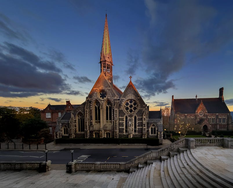 A historic stone church with intricate Gothic architecture and stained glass windows, featuring a tall spire reaching towards the evening sky with a mix of orange and blue hues; in the foreground, the