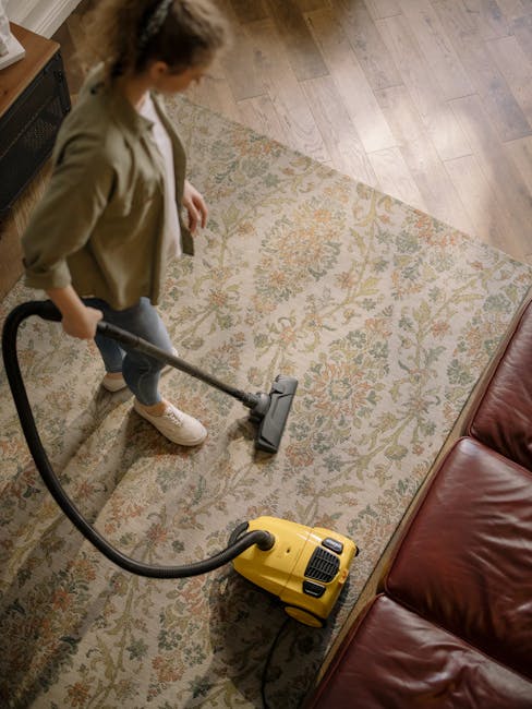 A person using a handheld vacuum cleaner connected to a yellow canister vacuum to deep clean a large, patterned carpet in a residential setting. The carpet features a floral design in muted tones of b