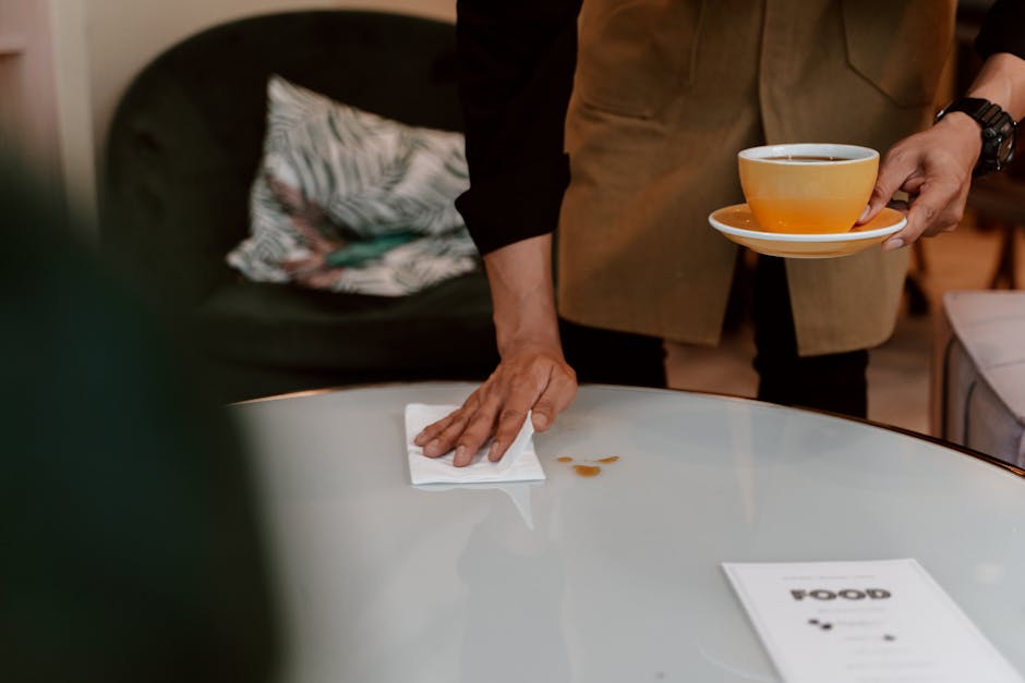 A person cleaning a white, smooth surface table with a white cloth, with a few small coffee stains visible on the table. The individual is holding a yellow cup and saucer in their left hand, while wiping the table with their right hand. The background includes a black sofa with a patterned cushion, and is lit with warm, ambient lighting. The scene depicts domestic cleaning and surface sanitisation, aligning with services offered by Carpet Cleaners Harrow for deep cleaning and stain removal in a home environment.