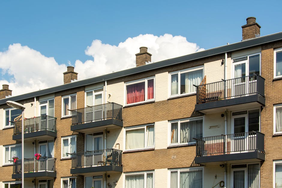 A multi-story residential building with a facade featuring a combination of brickwork and white panels, showcasing several balconies with black metal railings. The balconies are equipped with outdoor furniture such as chairs and potted plants, and have glass doors leading into the flats. The building's windows are framed in white, with some curtains visible inside. Bright daylight illuminates the building, with a partly cloudy sky in the background. The overall appearance is clean and well-maintained, reflecting professional property upkeep and care, consistent with the cleaning services offered by Carpet Cleaners Harrow at the End of tenancy cleaning South Harrow flats.