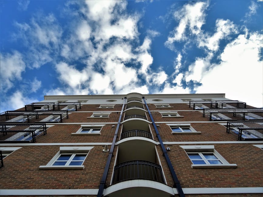 Low-angle view of a multi-story residential building with brick facade and white accents, featuring several black metal balconies. The building's windows have white frames and are evenly spaced along each floor. The scene is set against a partly cloudy sky with patches of blue visible through the white clouds. The exterior appears clean and well-maintained, with no visible dirt or debris, highlighting the pristine condition suitable for fostering hygiene in residential settings. This image visually supports the importance of surface cleaning and maintenance as part of professional domestic cleaning or end of tenancy cleaning services by Carpet Cleaners Harrow.