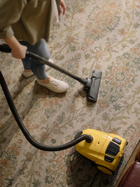 A person cleaning a patterned beige and green rug using a vacuum cleaner with a black wand attachment. The individual is dressed in light-colored shoes, blue jeans, and a beige jacket. The vacuum clea