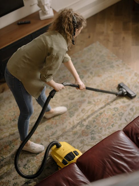 A person cleaning a patterned beige and green rug using a vacuum cleaner with a black wand attachment. The individual is dressed in light-colored shoes, blue jeans, and a beige jacket. The vacuum clea