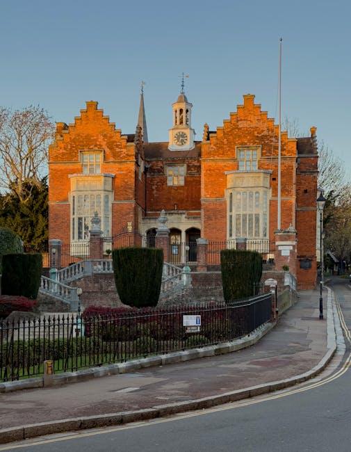 A historic red-brick building with a central clock tower and ornate architectural details, situated on a street corner. The building features large multi-pane windows, decorative stone accents, and a steeply pitched roof with small spires. In front of the structure, there is a well-maintained garden with neatly trimmed shrubs, a black wrought iron fence, and a paved sidewalk. The scene is illuminated by natural daylight, highlighting the building's historic charm and well-preserved façade. As part of professional cleaning services offered by Carpet Cleaners Harrow, such deep cleaning enhances the appearance and hygiene of residential and commercial properties in North Harrow estates and Harrow station areas.