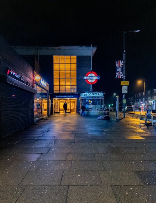 A person standing outdoors at a street crossing near South Harrow station, holding a large blue umbrella that obscures their upper body. The individual is dressed in dark clothing and carries a black bag. The background features a brick building with a sign indicating the station and traffic lights. The scene is overcast, suggesting rainy weather, highlighting the importance of surface cleaning and protection from weather elements. Carpet Cleaners Harrow offers professional cleaning services for such environments, ensuring surfaces remain clean, safe, and well-maintained.
