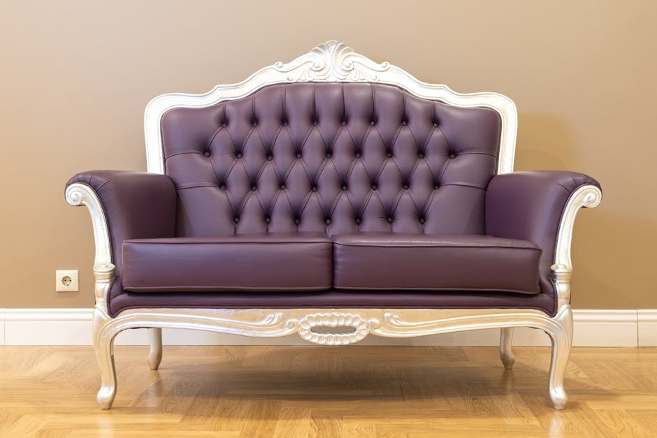 A vintage-style sofa with a white carved wooden frame and deep purple, tufted leather upholstery is positioned against a beige wall in a well-lit room. The sofa features elegant curved armrests, sturdy cabriole legs, and a high, elaborately decorated backrest, all appearing clean and polished. The room has a wood floor, and a white electrical outlet is visible near the left side of the sofa. This setting exemplifies surface cleaning and maintenance typical of domestic upholstery care provided by Carpet Cleaners Harrow, focusing on surface hygiene and deep cleaning for furniture.