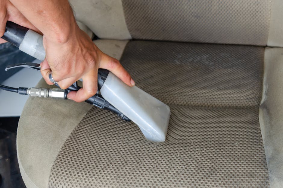 A close-up view of a person's hand operating a handheld upholstery cleaning tool on a beige fabric car seat. The tool is connected to a source of cleaning solution or suction device, and is used to deep clean and sanitise the fabric surface, which displays areas of visible dirt and stains. The upholstery has a textured, patterned surface with a mix of smooth and slightly worn sections. The room lighting highlights the contrast between the cleaned and uncleaned areas, demonstrating surface cleaning processes, and the scene is set in a vehicle interior. The image showcases professional upholstery cleaning, as provided by Carpet Cleaners Harrow, aimed at maintaining hygienic, fresh, and well-maintained domestic and commercial furniture surfaces.