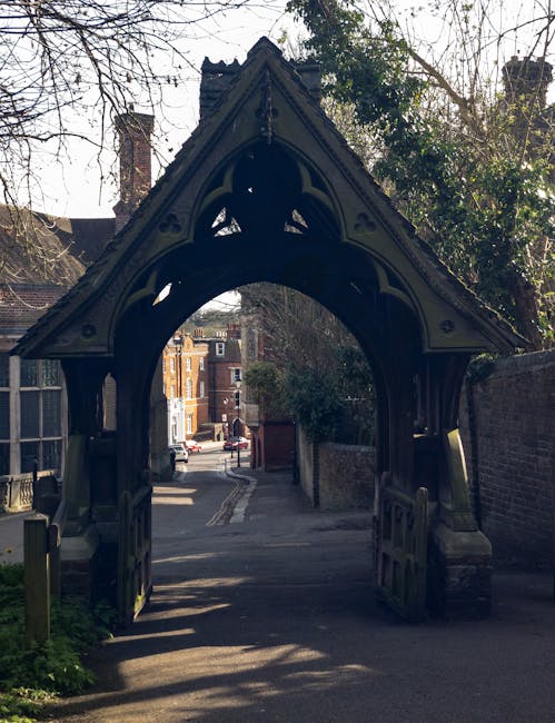 Photograph of an ornate, dark-colored wooden archway entrance leading into a residential street. The arch features intricate Gothic-style carvings and is supported by two sturdy posts. Beyond the arch, a narrow paved pathway extends into a neighborhood with brick buildings, cars parked along the street, and trees lining the roads. Natural daylight illuminates the scene, casting shadows on the ground. The area appears clean and well-maintained, reflecting a quiet suburban setting. This image is associated with Carpet Cleaners Harrow and relates to the topic of access problems for cleaning services in Harrow, highlighting the importance of navigating architectural features for service providers involved in surface cleaning, deep cleaning, and sanitisation within residential and commercial properties.